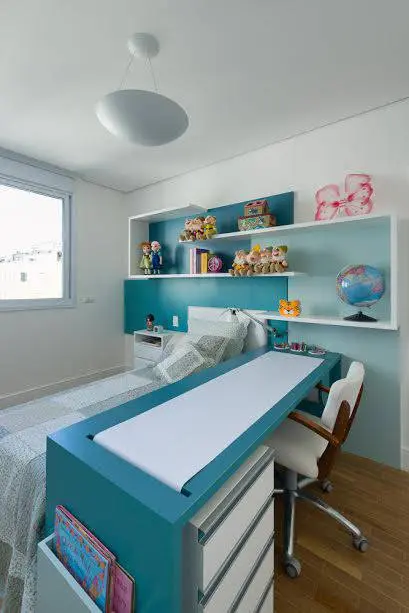 Bancadas de estudos azul petróleo e branca. Tem o formato de ilha e está em um quarto, ao lado da cama da moradora.