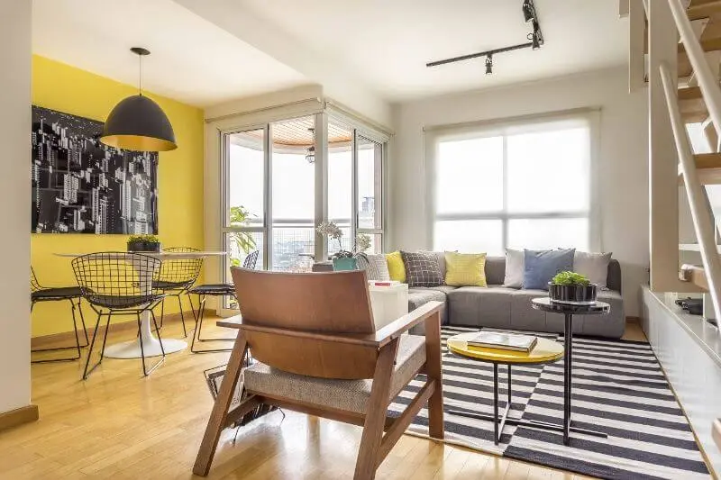 Sala integrada com parede amarela e sofá cinza e almofadas coloridas Projeto de Hildebrand Silva Arquitetura