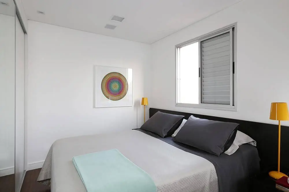 quarto de casal moderno - cama centralizada, com janela branca atrás e abajur amarelo ao lado