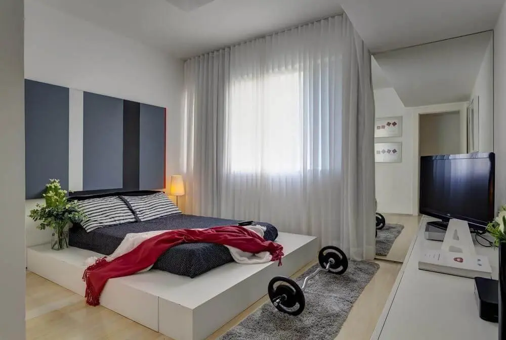 quarto de casal moderno - cama em cima de tapume em frente a um peso de musculação e televisão