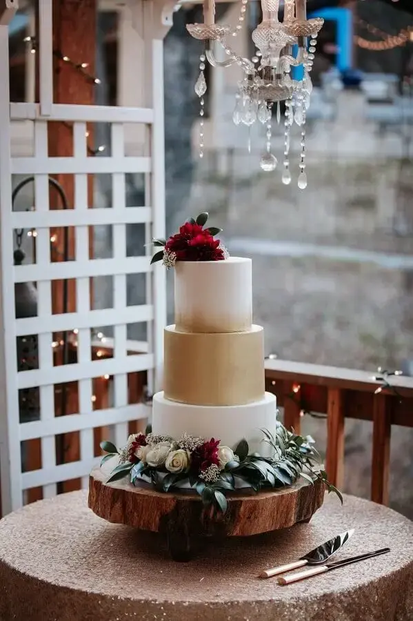 bolo de aniversário de casamento branco e dourado decorado com flores brancas e vinho Foto Assetproject