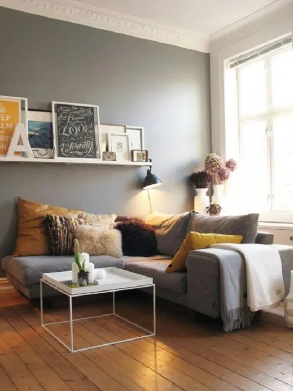 O sofá de canto cinza claro foi alinhado rente a parede e a janela do ambiente