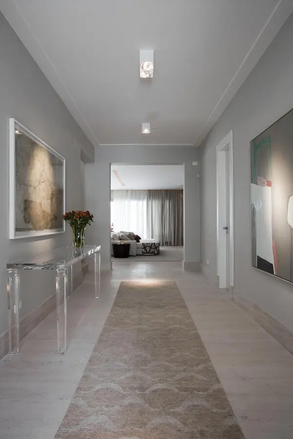 A luminária para corredor interno branca se conecta com o aparador acrílico e o tapete com estampa neutra