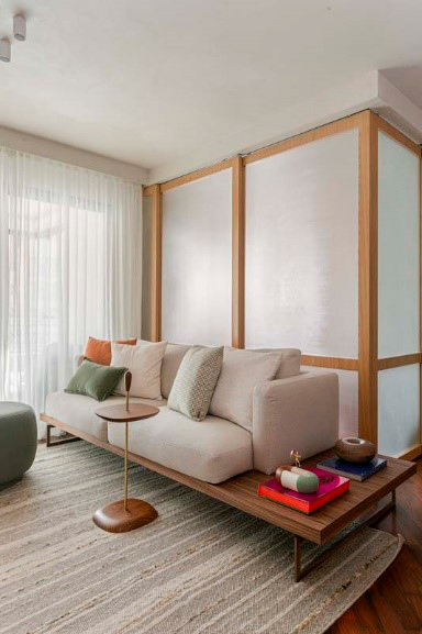 Sala com sofá branco sobre uma estrutura de madeira, no centro de uma sala integrada a outro ambiente, e uma porta de correr de vidro e madeira fechada.